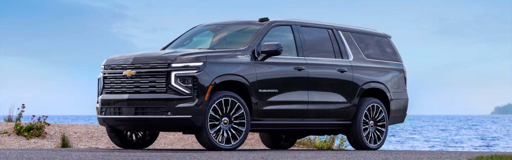 A sleek black 2025 Chevy Suburban SUV parked along the scenic shore of Houghton Lake, MI, on a sunny day. Gentle blue waves ripple under a vibrant blue sky, while to the left, a natural landscape of broken shells and native green plants adds a touch of rugged beauty.