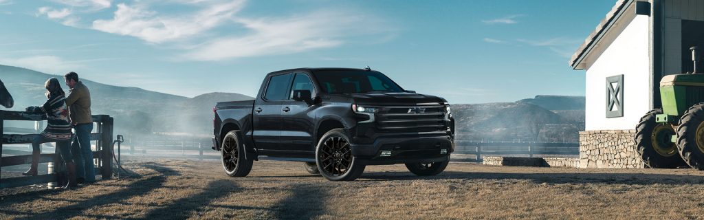 A sleek black 2025 Chevrolet Silverado 1500 parked on a dirt driveway at a farm near Houghton Lake, MI. To the right, a white farmhouse and a green tractor partially enter the frame. On the left, a man and woman lean over a wooden fence, chatting beside a horse. The backdrop features towering mountains beneath a bright blue sky on a sunny day.