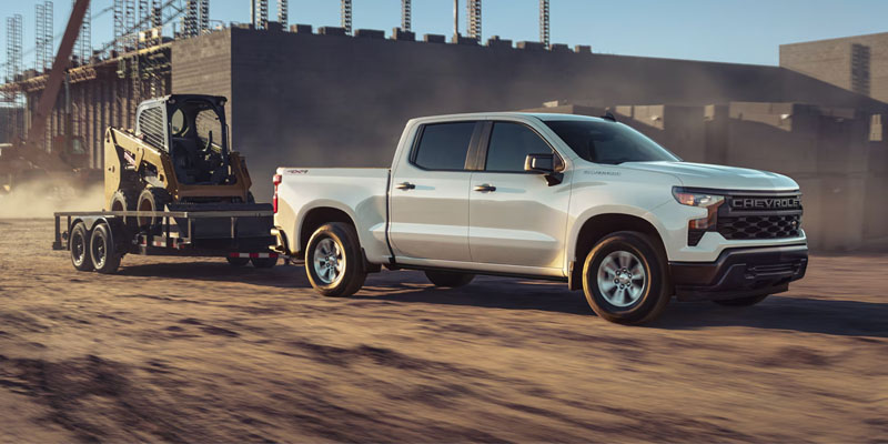A white Chevrolet Silverado 1500 truck towing a trailer with construction equipment in a desert construction site.